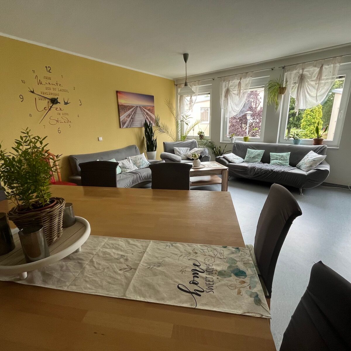 Vom Holztisch aus kann man Sitzmöglichkeiten sehen und ein große Wanduhr.