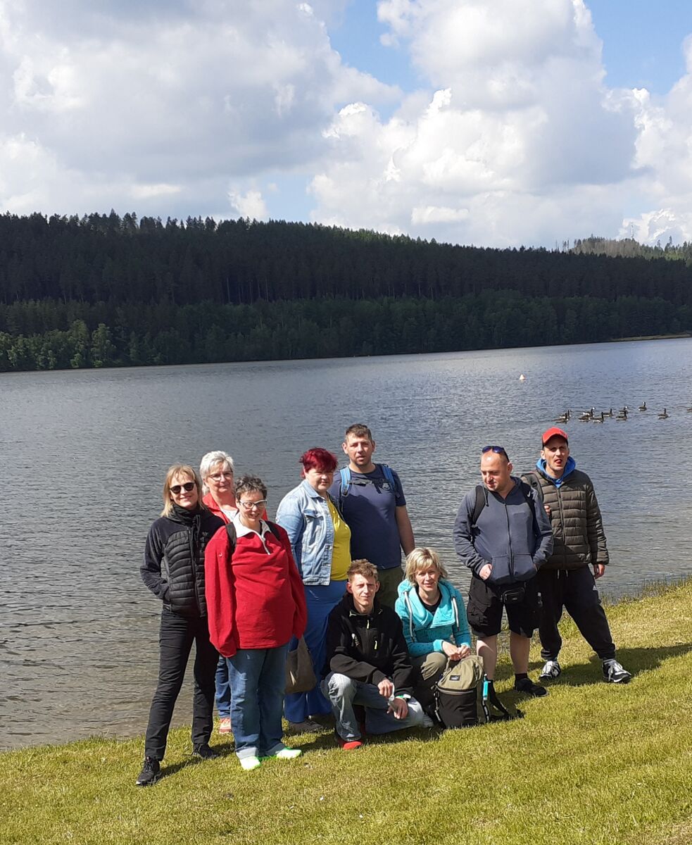 Neun Menschen machen einen Gruppenfoto, hinter ihnen ist ein See und Nadelwald. Auf dem Wasser sind Enten zu sehen.